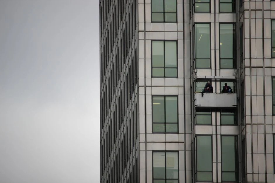 Two workers clean windows on a modern glass building facade using a suspended platform.